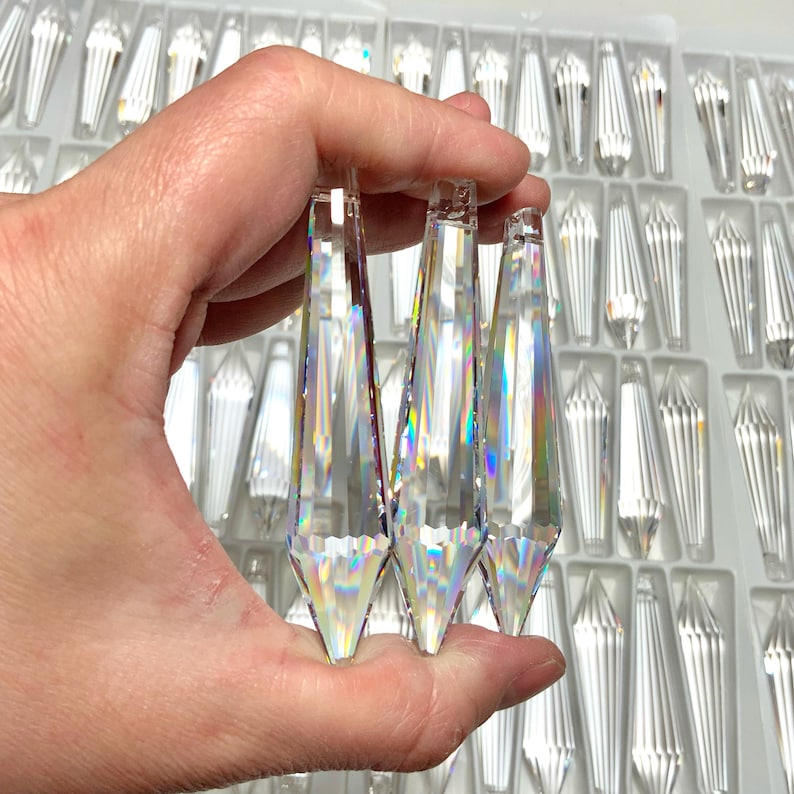 May include: Three clear crystal teardrop-shaped prisms with iridescent rainbow facets. The prisms are held in a hand against a background of many similar prisms.
