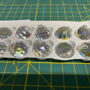 K&ouml;nnte beinhalten: Eine Packung mit 10 klaren Kristall-Prismenanh&auml;ngern mit schillernden Regenbogen-Facetten. Die Anh&auml;nger sind in einer wei&szlig;en Kunststoffschale mit einzelnen F&auml;chern angeordnet.