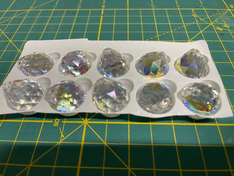K&ouml;nnte beinhalten: Eine Packung mit 10 klaren Kristall-Prismenanh&auml;ngern mit schillernden Regenbogen-Facetten. Die Anh&auml;nger sind in einer wei&szlig;en Kunststoffschale mit einzelnen F&auml;chern angeordnet.
