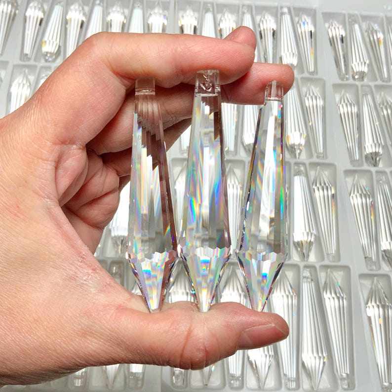 May include: Three clear crystal teardrop prisms of varying sizes, held in a hand. The prisms are faceted and reflect light, creating a rainbow effect.