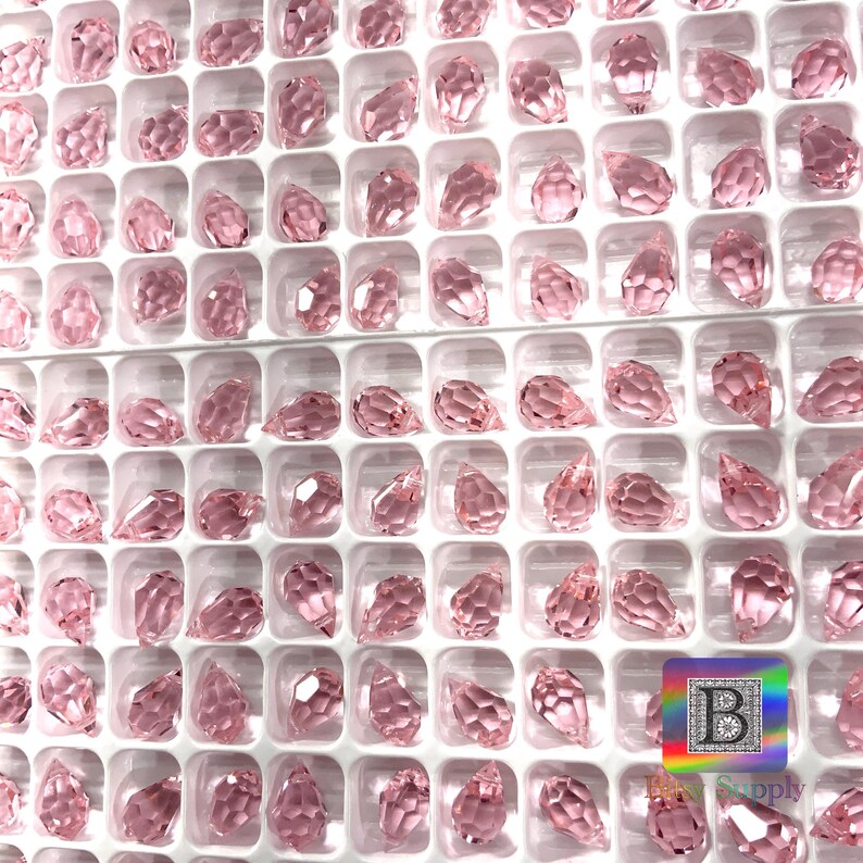 May include: A close-up view of a white plastic tray filled with pink crystal teardrop beads. The beads are arranged in rows and columns, and they are all the same size and shape.