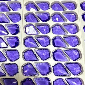 May include: Rows of faceted, irregular-shaped, purple glass stones are arranged in a white tray. The stones have a glossy, reflective surface, and the tray is divided into individual compartments for each stone. The overall effect is a display of sparkling, decorative elements.