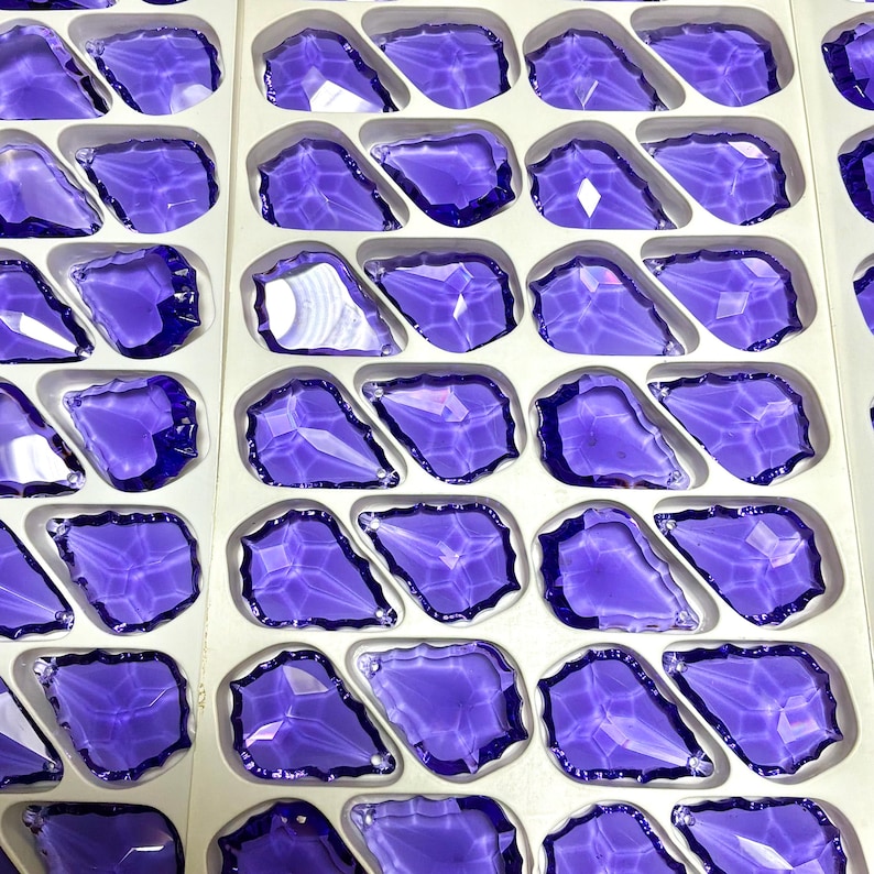 May include: Rows of faceted, irregular-shaped, purple glass stones are arranged in a white tray. The stones have a glossy, reflective surface, and the tray is divided into individual compartments for each stone. The overall effect is a display of sparkling, decorative elements.
