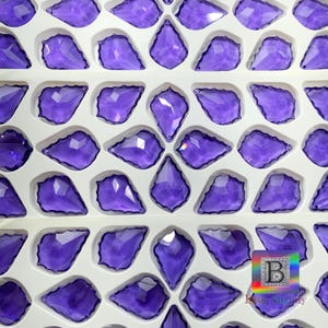 Peut inclure: Collection de pendentifs en cristal violets, facett&eacute;s et en forme de goutte d'eau, dispos&eacute;s dans un plateau blanc. Les cristaux sont d'un violet profond et r&eacute;fl&eacute;chissent la lumi&egrave;re. Les pendentifs sont pr&eacute;sent&eacute;s en rang&eacute;es.