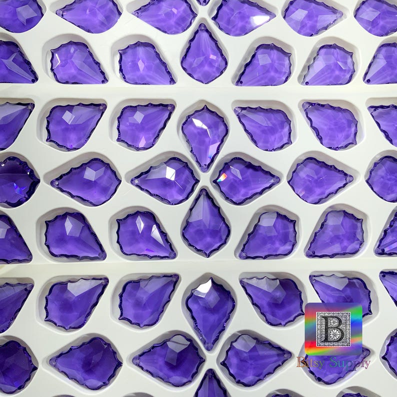 Peut inclure: Collection de pendentifs en cristal violets, facett&eacute;s et en forme de goutte d'eau, dispos&eacute;s dans un plateau blanc. Les cristaux sont d'un violet profond et r&eacute;fl&eacute;chissent la lumi&egrave;re. Les pendentifs sont pr&eacute;sent&eacute;s en rang&eacute;es.