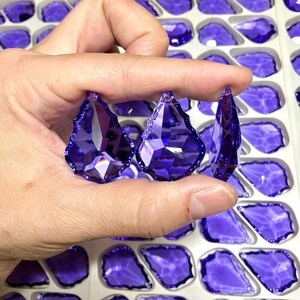 May include: Three faceted, teardrop-shaped, purple crystal pendants are held between fingers. The crystals have a pointed top and a jagged edge. The background shows more purple crystals in a white tray.