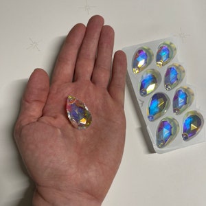 May include: A close-up of iridescent teardrop-shaped crystal embellishments. The crystals are arranged in a clear plastic packaging. One crystal is being held in a hand.