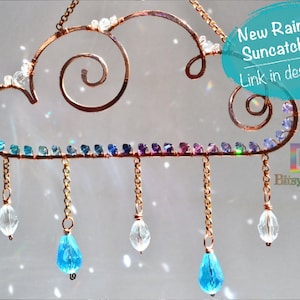 May include: A copper wire suncatcher shaped like a cloud with a swirl design. The cloud has a rainbow of small blue, purple, and green beads around the edge.  The cloud has five chains hanging from it, each with a crystal bead at the end.  The crystal beads are clear and blue.  The text "New Raincloud Suncatcher Kit Link in description!" is in a blue circle on the right side of the image.