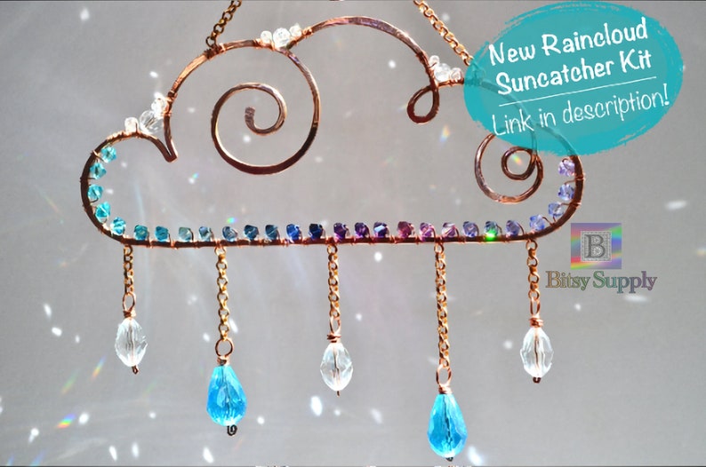 May include: A copper wire suncatcher shaped like a cloud with a swirl design. The cloud has a rainbow of small blue, purple, and green beads around the edge.  The cloud has five chains hanging from it, each with a crystal bead at the end.  The crystal beads are clear and blue.  The text "New Raincloud Suncatcher Kit Link in description!" is in a blue circle on the right side of the image.