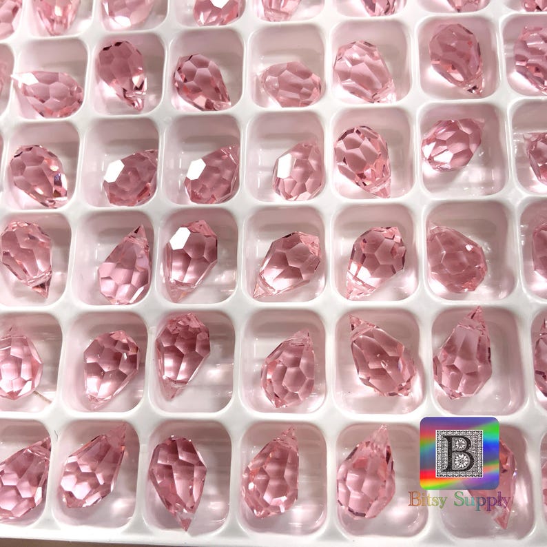 May include: A close-up view of a container filled with pink crystal teardrop shaped beads. The beads are arranged in rows and columns, and they are all the same size and shape.