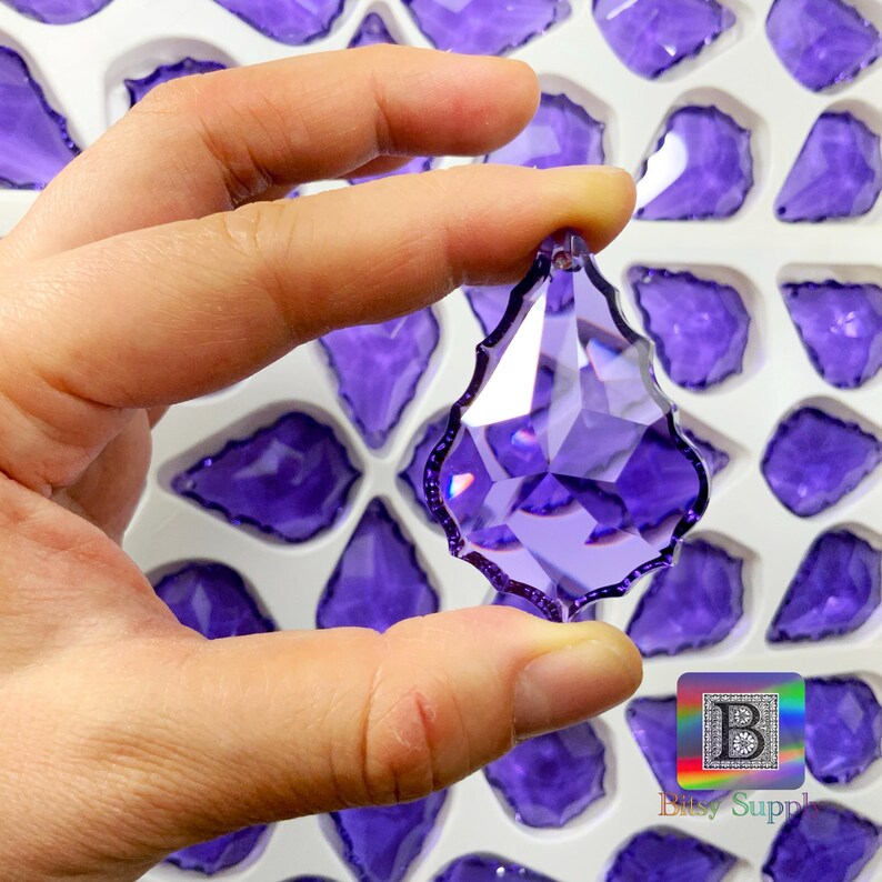 Peut inclure: Un pendentif en cristal violet en forme de goutte d'eau facett&eacute;e est tenu entre les doigts. Le pendentif a un bord d&eacute;coratif festonn&eacute; et est entour&eacute; de cristaux similaires dans un plateau blanc. Les cristaux sont d'un violet profond et vibrant.