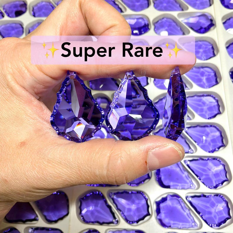 May include: Three faceted, teardrop-shaped, purple crystals are held in a hand, with the text "Super Rare" across the top. The crystals are surrounded by other purple crystals in a tray. The crystals have a high-gloss finish.