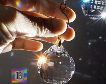 May include: A clear, faceted crystal ball pendant with a gold-colored cap is held by a hand. The crystal reflects light, creating a rainbow effect. A second crystal ball is visible in the background. The logo "Bitsy Supply" is in the lower left corner.