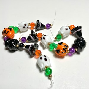 May include: Halloween-themed beaded strands with glass beads shaped like ghosts, witch hats, and jack-o'-lanterns. The beads are in orange, black, white, green, purple, and gold. The strands are strung together with a thin, clear string.