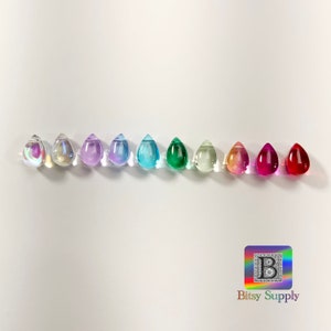 May include: Ten teardrop-shaped glass beads in various colors, including iridescent, lavender, light blue, teal, green, clear, orange, red, and pink. The beads are arranged in a row on a white surface.