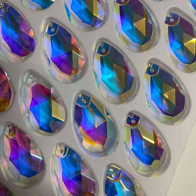 May include: A sheet of iridescent teardrop-shaped glass rhinestones in various shades of blue, purple, and gold. The rhinestones are arranged in a grid pattern on a white background.