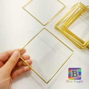 Puede incluir: Aro de metal cuadrado dorado, perfecto para crear joyas o decoración para el hogar. Los aros se muestran apilados sobre una superficie blanca.