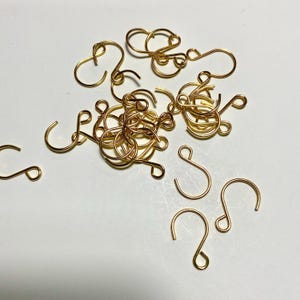 May include: A collection of gold-colored earring hooks and decorative wire components. The hooks are in various shapes, including curved and looped designs. The components are scattered on a white surface.