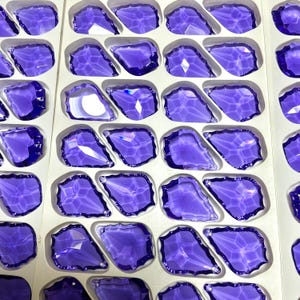 May include: Rows of faceted, purple glass pieces, each with a unique, irregular shape, are arranged in a white tray. The pieces have a glossy, reflective surface, catching the light and creating a sparkling effect. The overall theme is decorative glass elements.
