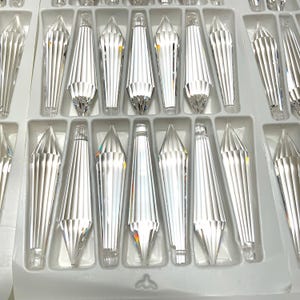 May include: A close-up view of a tray of clear crystal teardrop-shaped pendants. The pendants are arranged in rows and are all the same size and shape. The tray is white and has a textured surface.