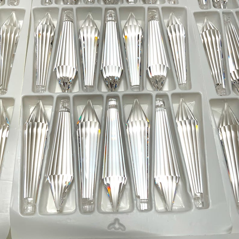 May include: A close-up view of a tray of clear crystal teardrop-shaped pendants. The pendants are arranged in rows and are all the same size and shape. The tray is white and has a textured surface.