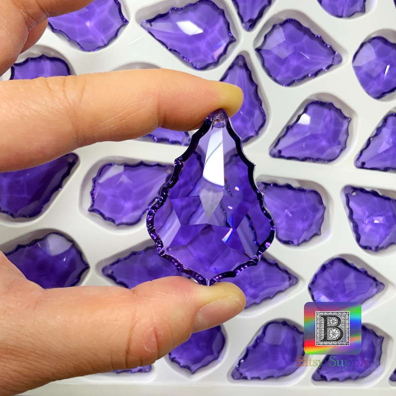 Peut inclure: Un pendentif en cristal violet en forme de goutte d'eau facett&eacute;e est tenu entre les doigts. Le pendentif a un bord festonn&eacute; et est entour&eacute; d'autres cristaux similaires dans un plateau blanc. Les cristaux sont d'un violet profond et transparent.
