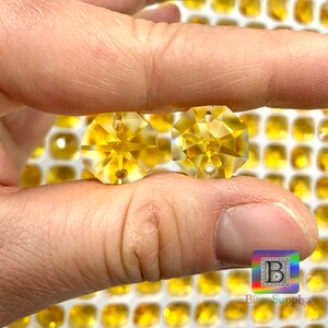 Puede incluir: Dos cuentas de cristal octogonales transparentes de color amarillo con un agujero en el centro. Las cuentas se sostienen en una mano sobre un fondo de muchas cuentas similares.
