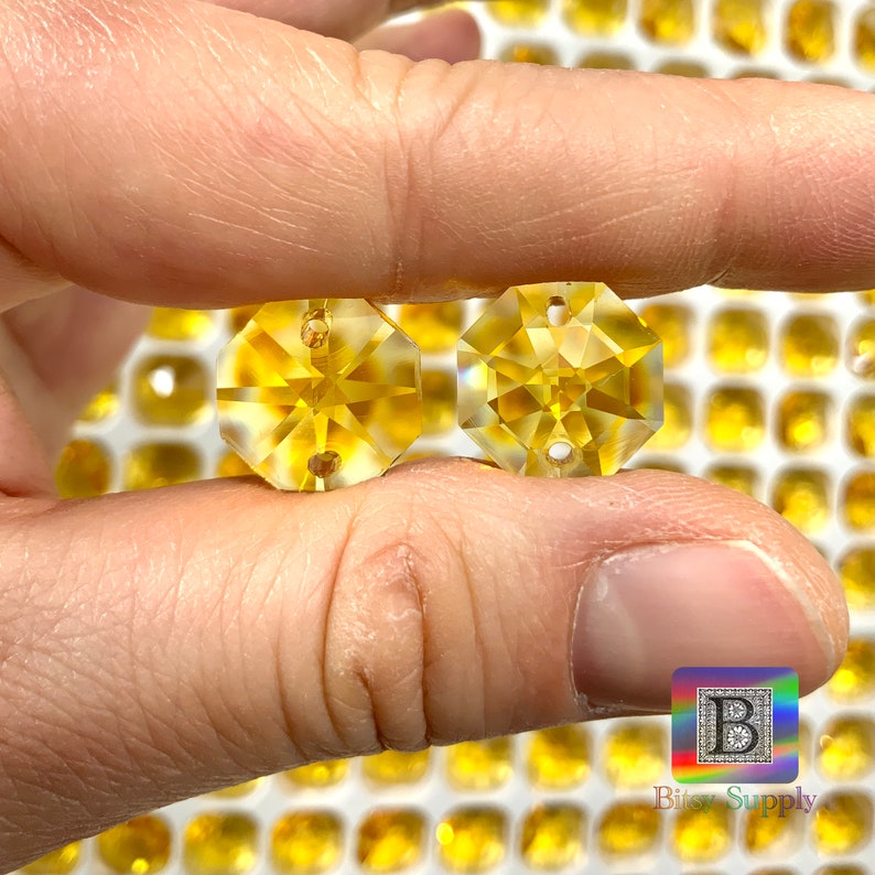 Puede incluir: Dos cuentas de cristal octogonales transparentes de color amarillo con un agujero en el centro. Las cuentas se sostienen en una mano sobre un fondo de muchas cuentas similares.