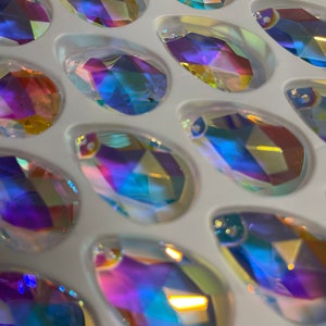 May include: A close-up of a tray of iridescent teardrop-shaped glass crystals. The crystals are arranged in rows and are all different shades of blue, purple, and green.