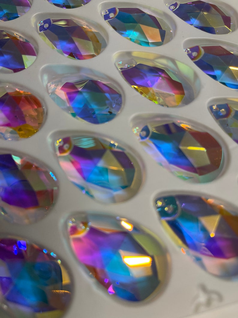 May include: A close-up of a tray of iridescent teardrop-shaped glass crystals. The crystals are arranged in rows and are all different shades of blue, purple, and green.