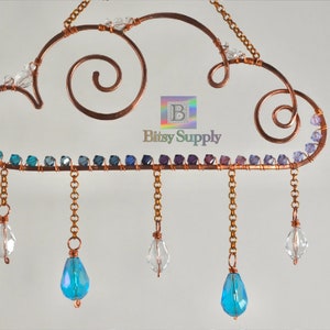May include: A copper wire cloud with a gradient of blue and purple beads and copper chains with crystal and blue teardrop beads hanging from it. The cloud has two copper wire swirls on top. The text "Bitsy Supply" is visible below the cloud.