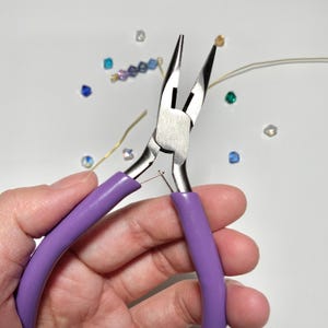 May include: Close-up of jewelry pliers with purple handles, used for crafting. The pliers are silver and are holding a small piece of wire. Scattered around are small, colorful beads and gold wire, suggesting jewelry making.