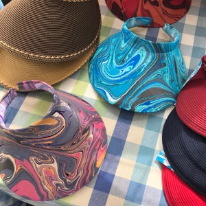 May include: Assortment of sun visors with marbled patterns in shades of blue, pink, and gray. The visors are made of fabric and have a wide brim.
