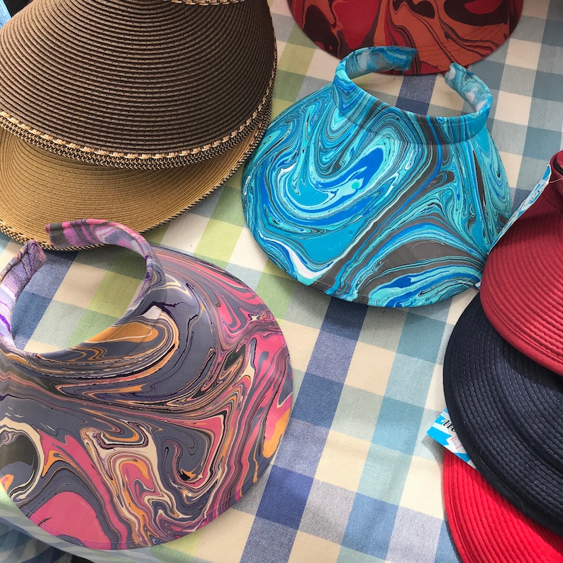 May include: Assortment of sun visors with marbled patterns in shades of blue, pink, and gray. The visors are made of fabric and have a wide brim.