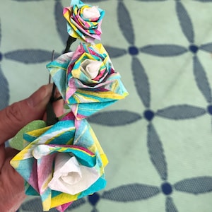 May include: Three paper roses in shades of blue, pink, and yellow with white centers. The roses are arranged in a cascading pattern and are attached to a stem.