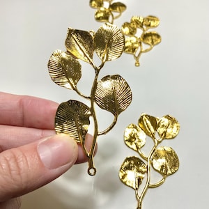 Könnte beinhalten: Goldfarbene Metallblattzweig-Anhänger. Die Anhänger haben detaillierte Adern auf den Blättern und eine Schlaufe zum Befestigen an Schmuck oder anderen Bastelarbeiten. Mehrere Anhänger sind vor weißem Hintergrund zu sehen.