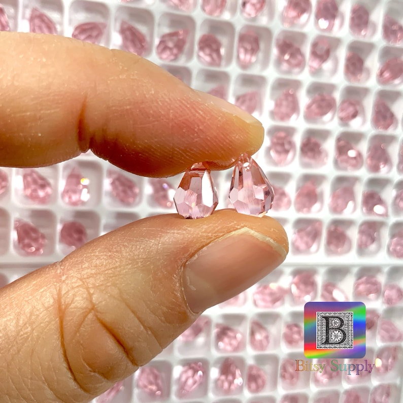 May include: A close-up of two pink crystal teardrop beads. The beads are faceted and have a shiny finish. The beads are held in a hand against a background of many other pink crystal beads.