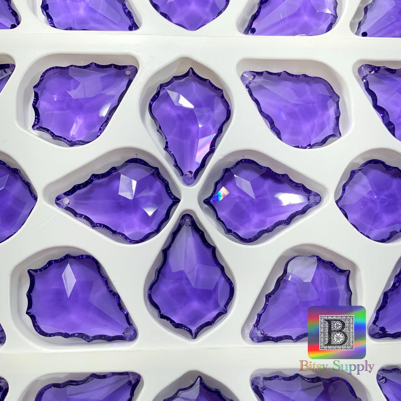 Peut inclure: Gros plan d'un plateau rempli de plusieurs pendentifs en cristal violets en forme de goutte d'eau. Chaque pendentif a un bord festonn&eacute; et est plac&eacute; dans un compartiment blanc. Les cristaux sont d'une teinte violette vibrante.