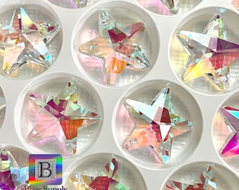 May include: A close-up of a white plastic container filled with iridescent star-shaped crystal embellishments. The container has a grid pattern and the stars are arranged in rows.