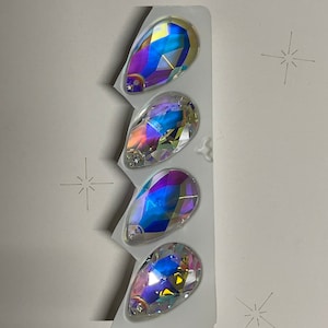 May include: Four iridescent teardrop-shaped glass rhinestones in a clear plastic packaging. The rhinestones have a rainbow effect with shades of blue, purple, and green.