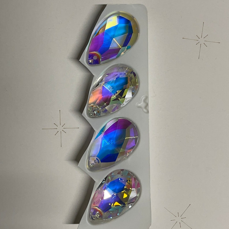 May include: Four iridescent teardrop-shaped glass rhinestones in a clear plastic packaging. The rhinestones have a rainbow effect with shades of blue, purple, and green.