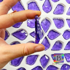 Peut inclure: Gros plan sur un cristal violet en forme de goutte d'eau facett&eacute;, tenu entre les doigts. Le cristal est d'un violet profond et a une surface textur&eacute;e. D'autres cristaux similaires sont dispos&eacute;s dans un plateau blanc. Le logo Bitsy Supply est en bas &agrave; droite.