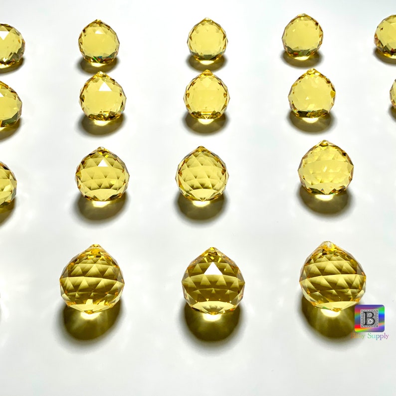 May include: A collection of 15 yellow crystal ball prisms, each with a faceted design, arranged in rows on a white surface.