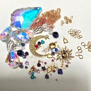 May include: A collection of celestial-themed jewelry-making supplies. Includes iridescent crystals, gold-colored charms of a crescent moon, sun, stars, and planets, along with gold chain and jump rings. Assorted colorful beads are also present.
