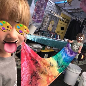 May include: Two children are painting a colorful scarf with rainbow colors. The scarf is made of fabric and has a marbled pattern. The children are wearing paint palettes on their eyes. The sign behind them says "Paint Your Own Scarf".