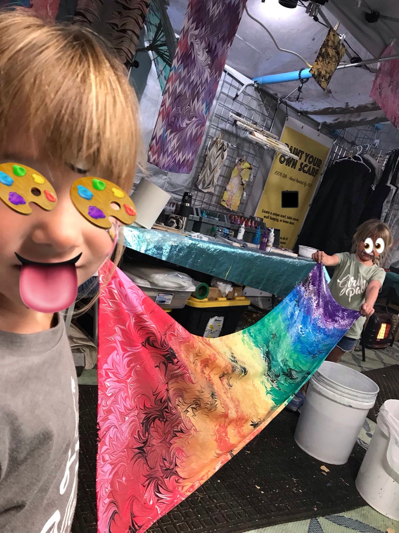 May include: Two children are painting a colorful scarf with rainbow colors. The scarf is made of fabric and has a marbled pattern. The children are wearing paint palettes on their eyes. The sign behind them says "Paint Your Own Scarf".