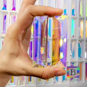 Peut inclure: Deux pendentifs en cristal prismatique iris&eacute;s, chacun de forme rectangulaire et &agrave; sommet pointu, sont tenus dans une main. Les prismes sont transparents et refl&egrave;tent des couleurs arc-en-ciel.