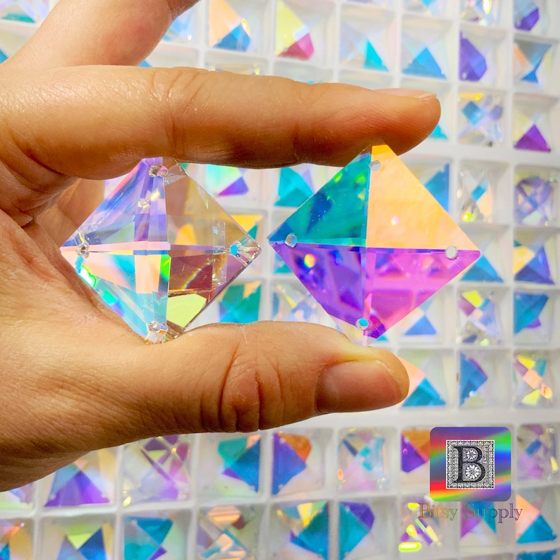 K&ouml;nnte beinhalten: Zwei regenbogenfarbene, quadratische Kristall-Strasssteine mit einem Regenbogeneffekt. Die Strasssteine werden in einer Hand gehalten.