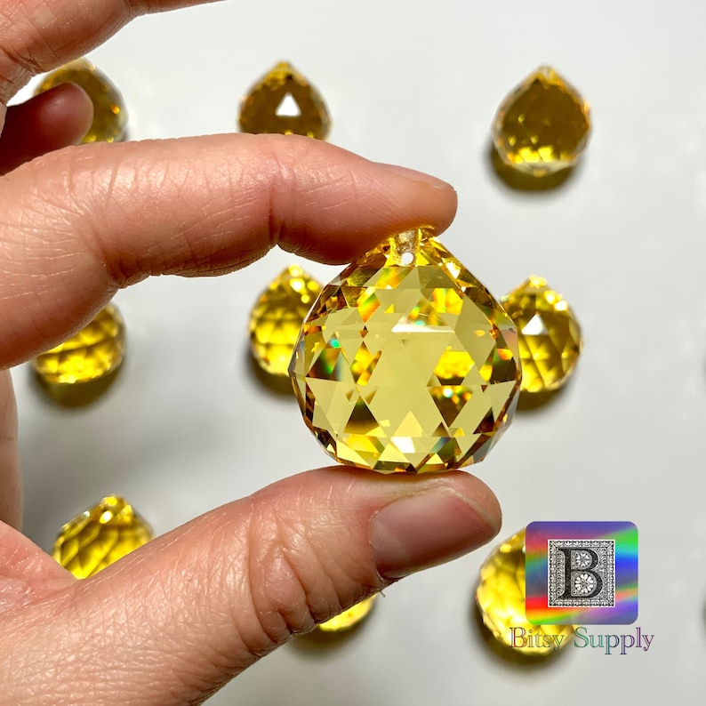 May include: A close-up of a hand holding a yellow crystal ball with a hole at the top. The crystal ball is faceted and reflects light. There are other yellow crystal balls in the background.