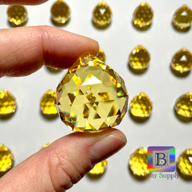 May include: A close-up of a yellow crystal ball ornament with many facets. The ornament is being held by a hand. The background is white.